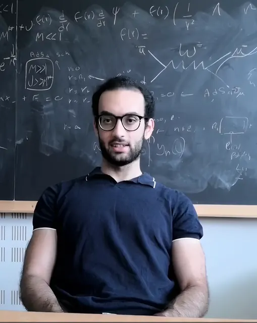 Ahmed speaking in front of a blackboard covered in equations and a Feynman diagram